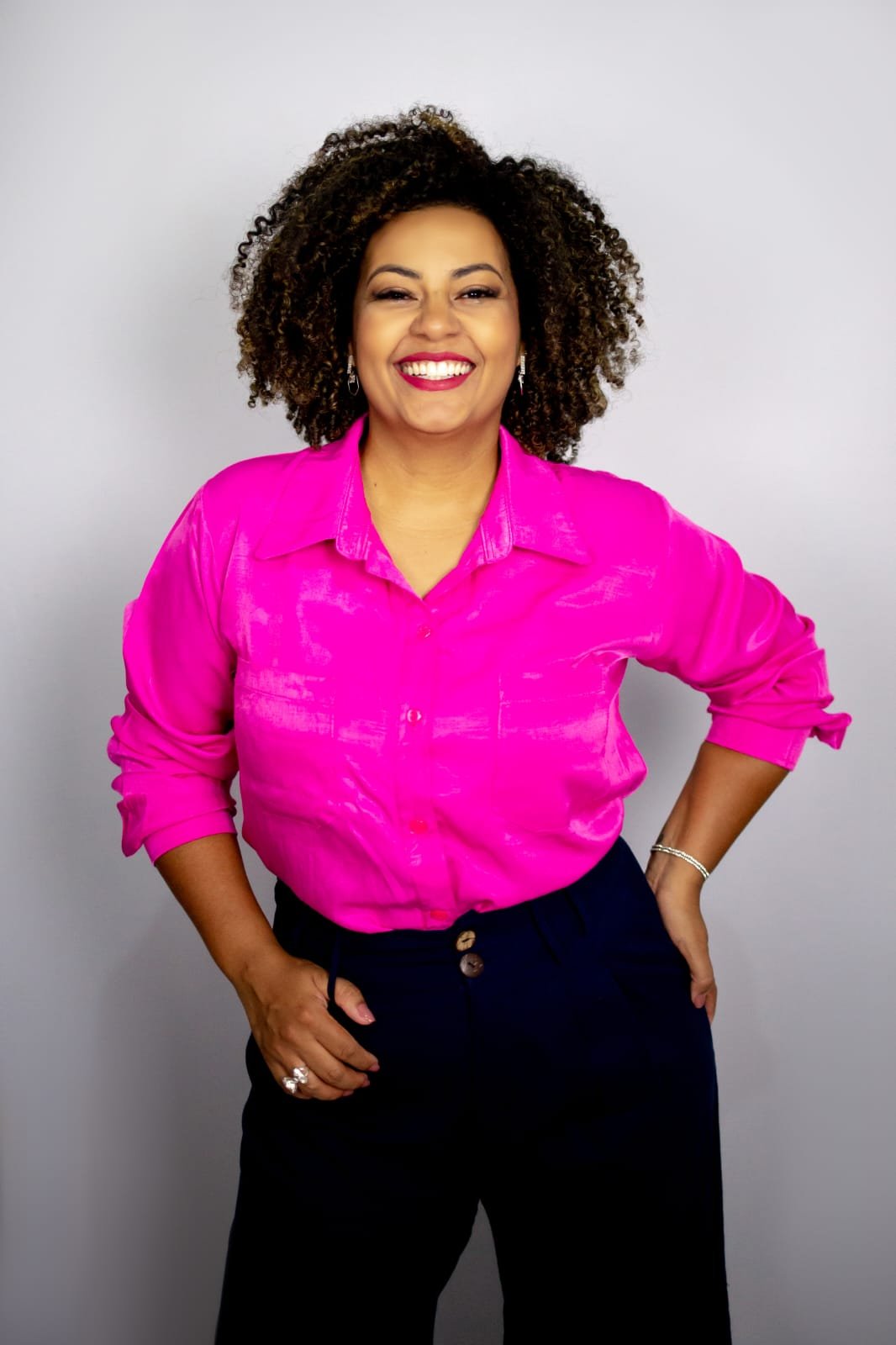 Anna Tavares, a musa do studio, de camisa rosa e mão na cintura, sorrindo pra quem chega